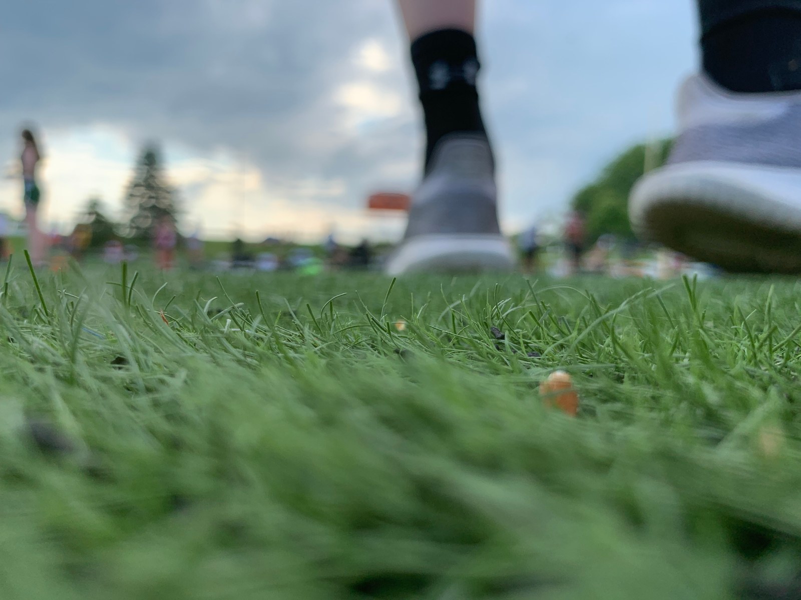 Green grass in focus with several people blurred beneath a cloudy sky