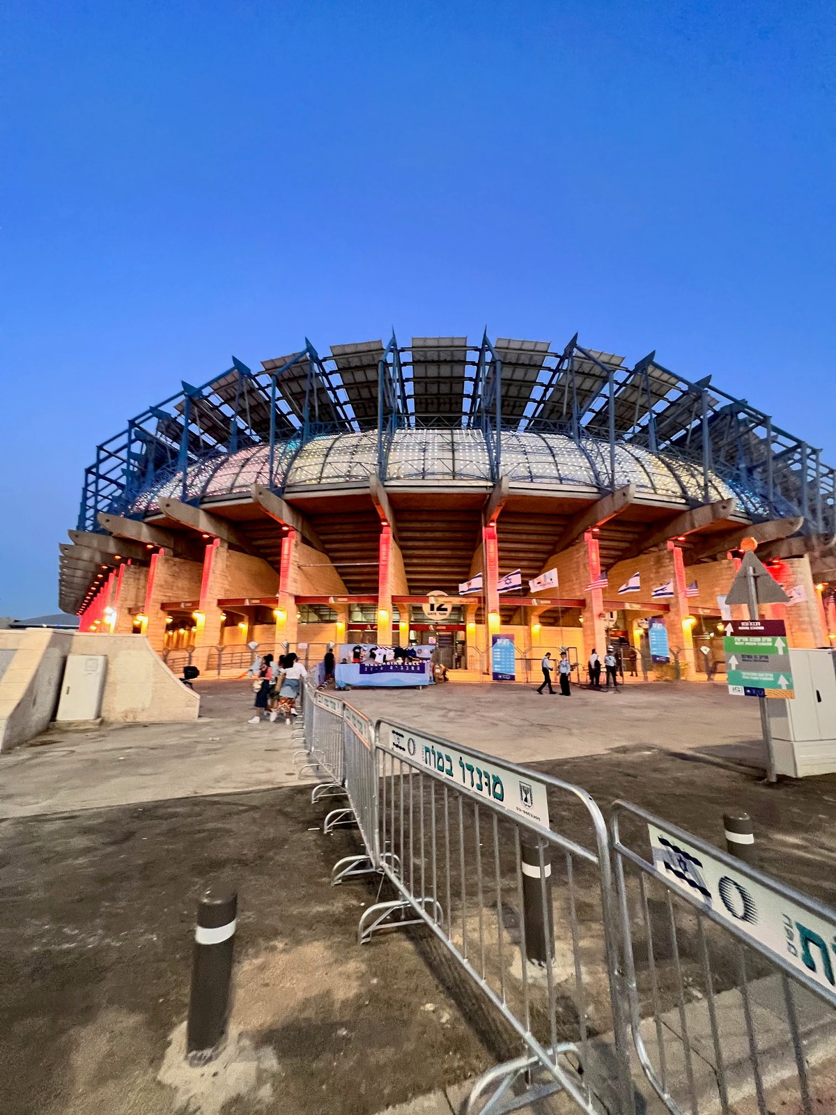 Stadium entrance lit red at dusk with Israeli flags, barriers, staff, and visitors
