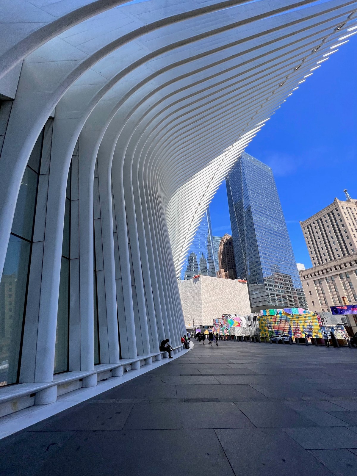 White ribbed architectural fins curving over a plaza beside glass-and-stone skyscrapers