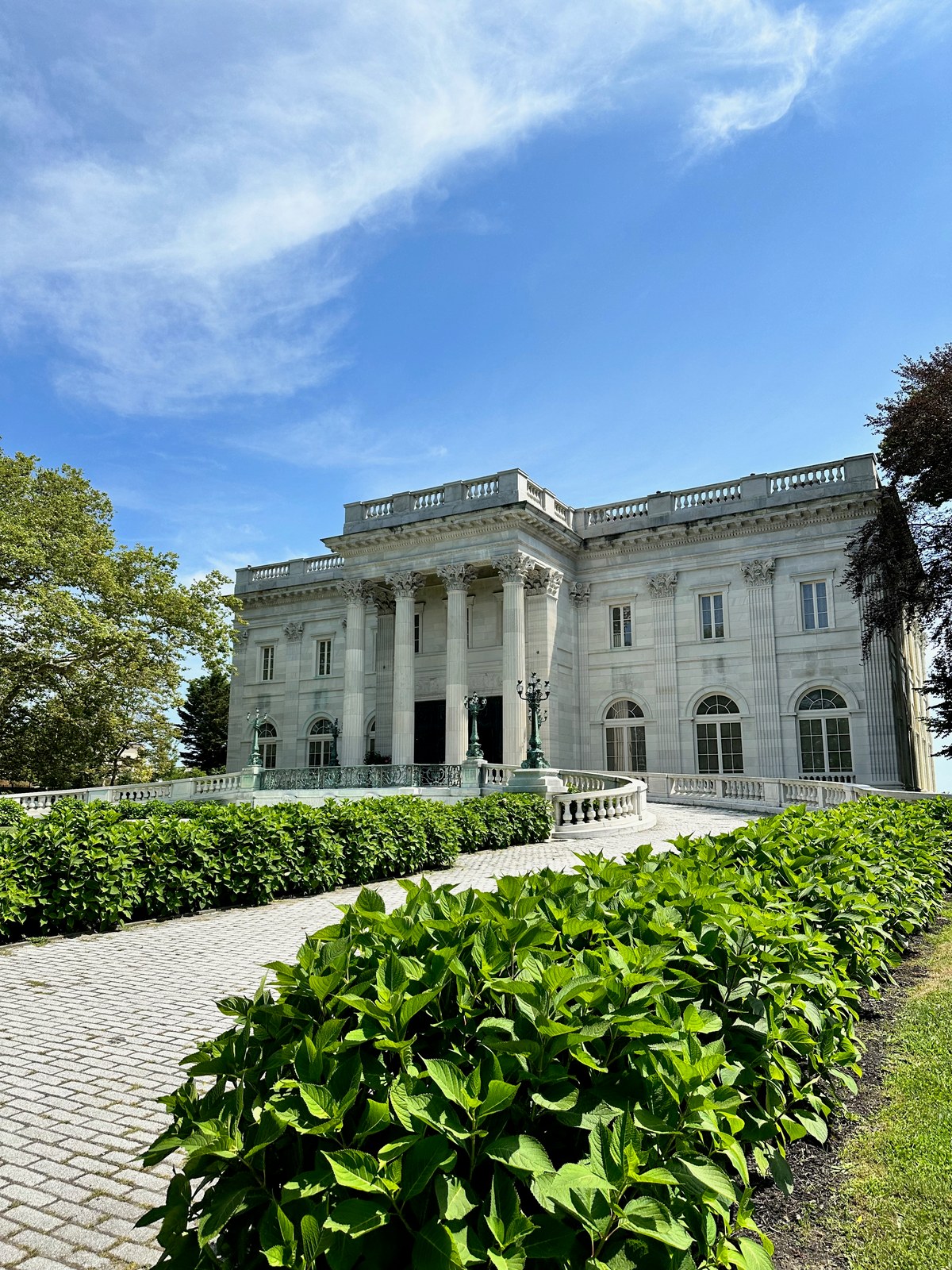 Grand white neoclassical mansion with Corinthian columns, balustrades, and surrounding garden beds