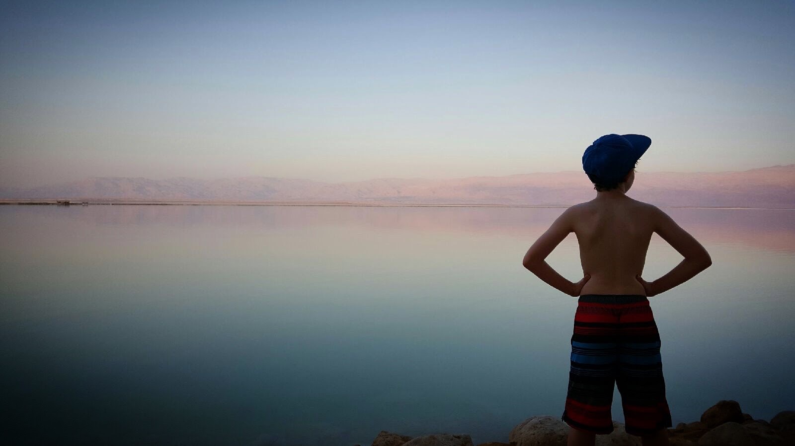 Child in striped swim trunks standing on rocks before calm water and distant mountains at dusk