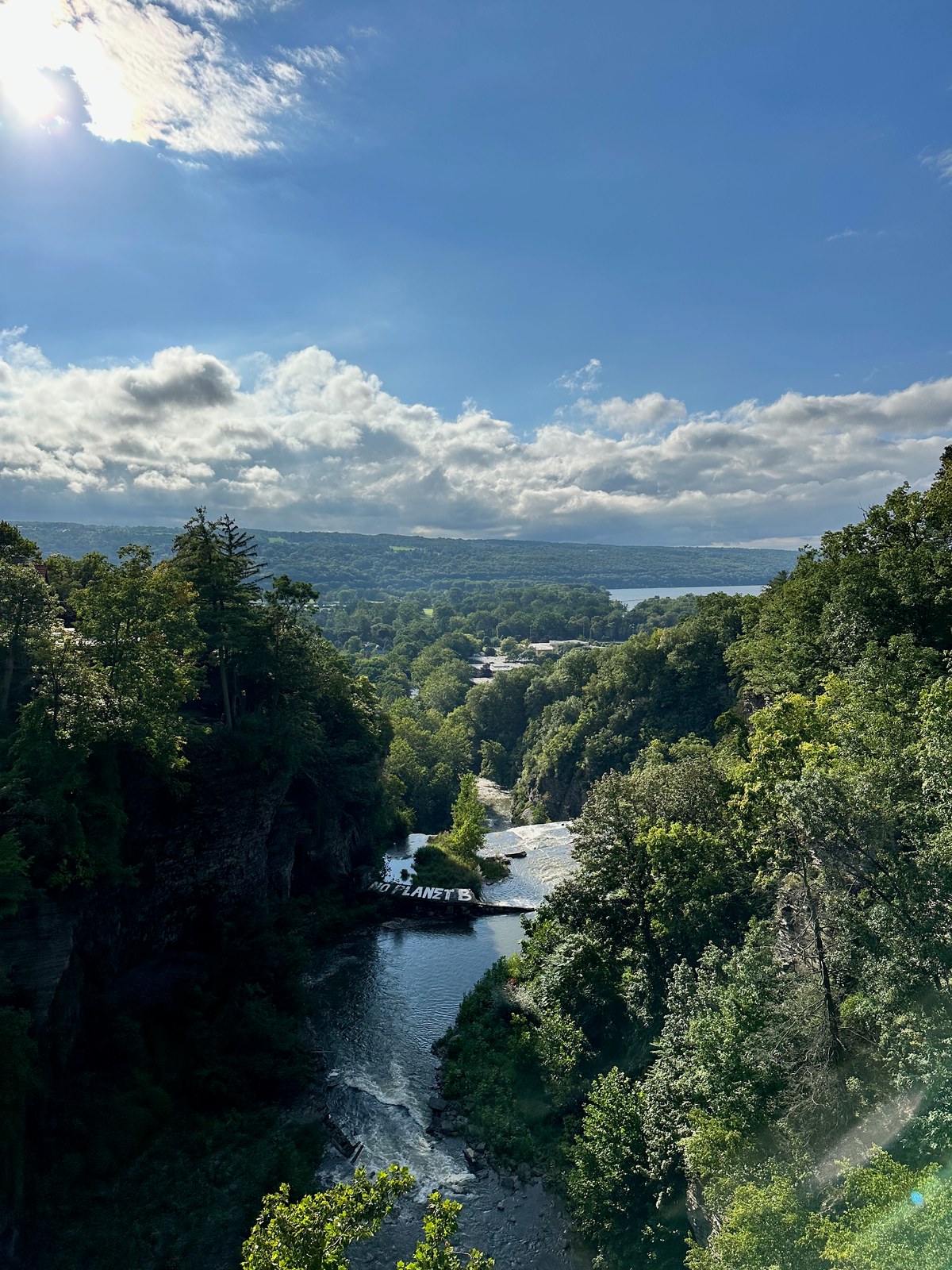 Tree-lined gorge with a cascading river, distant lake, and cloud-filled sky