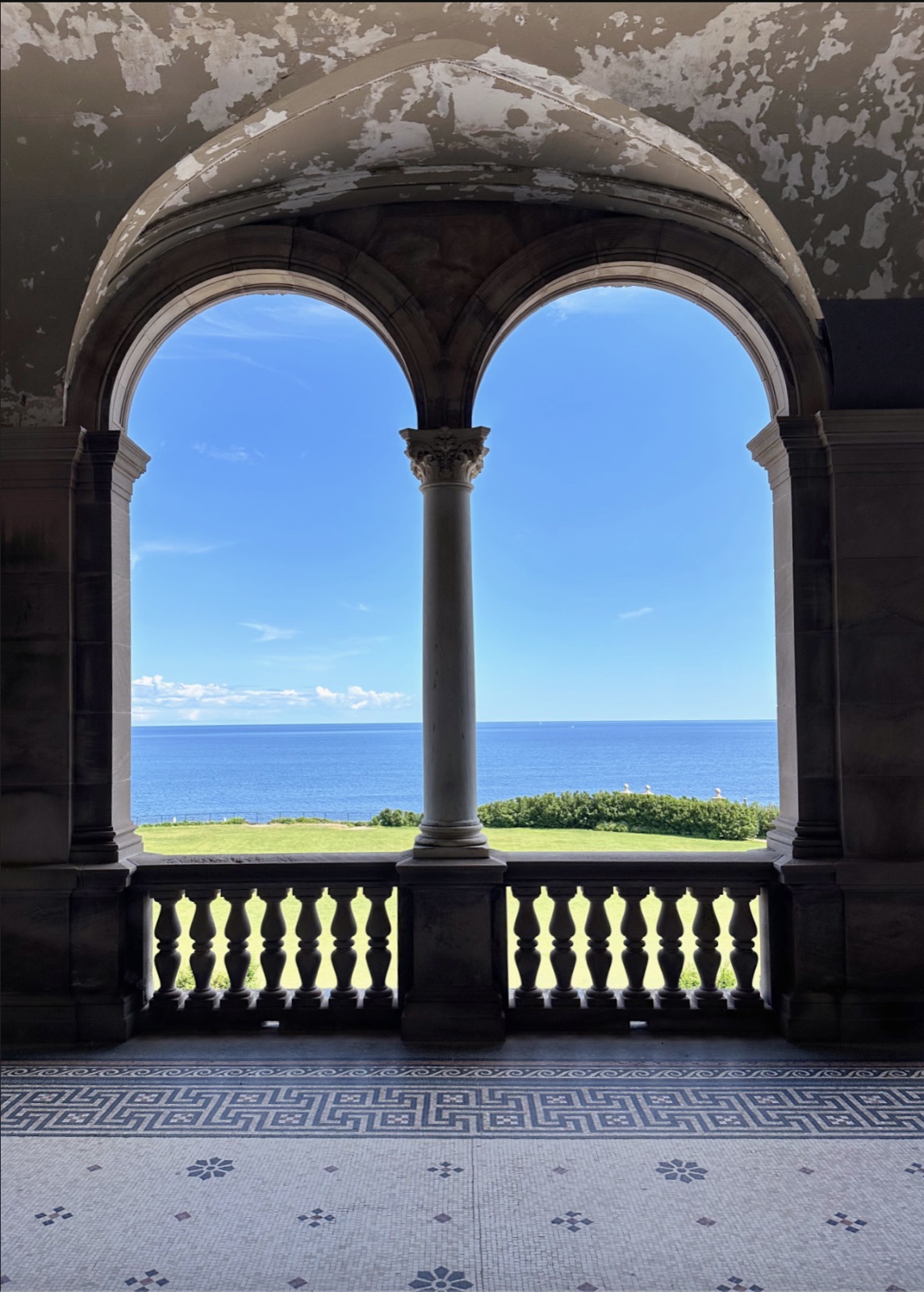 Twin stone arches and a central column framing a lawn and blue ocean above a patterned mosaic floor