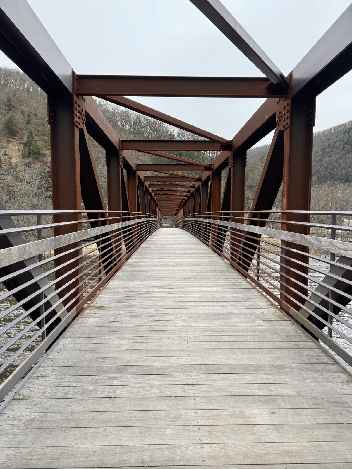 Empty wooden-deck steel truss bridge spanning a river through a leafless wooded valley