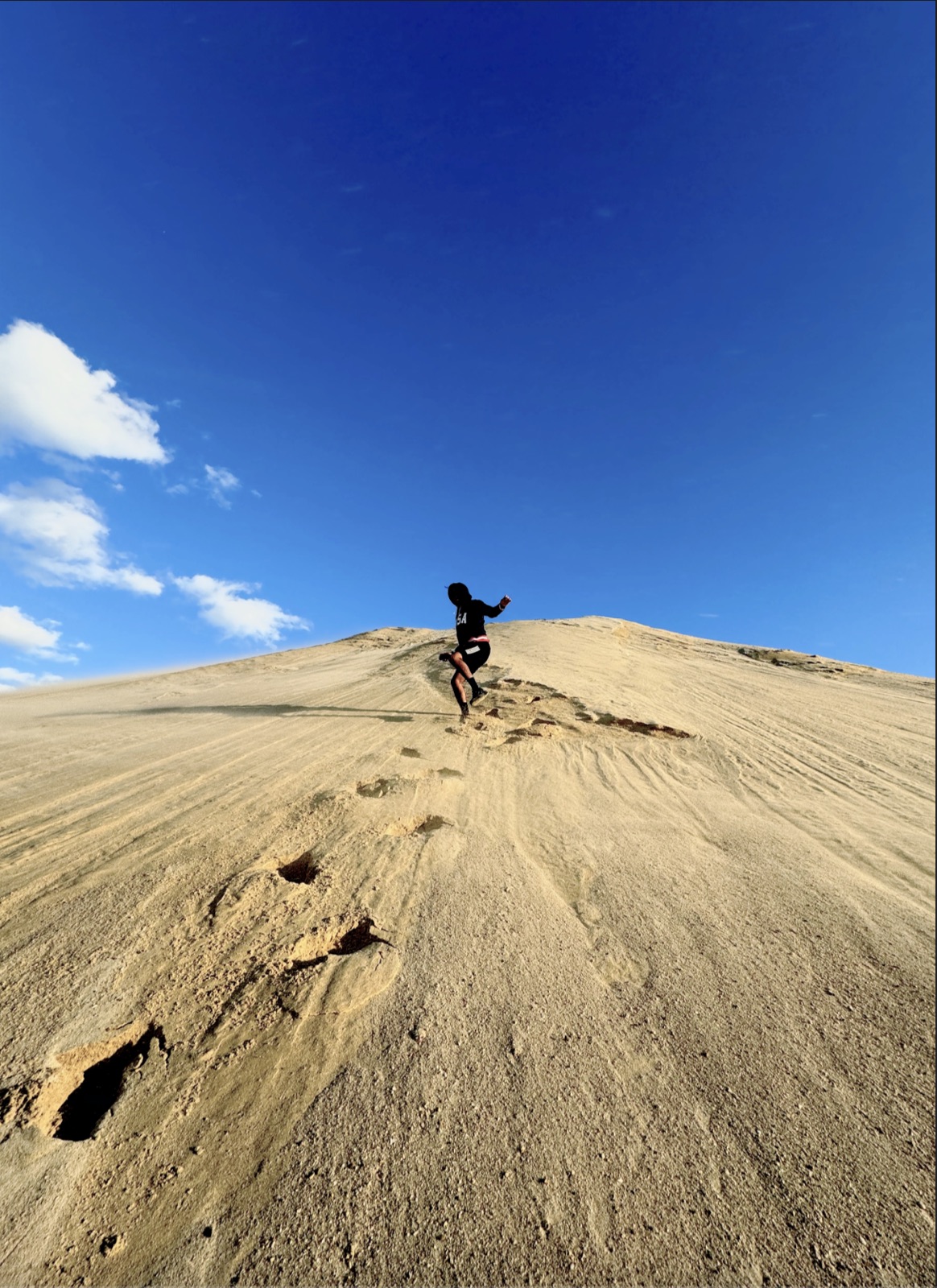 Person in black running up a steep sand dune along a trail of footprints