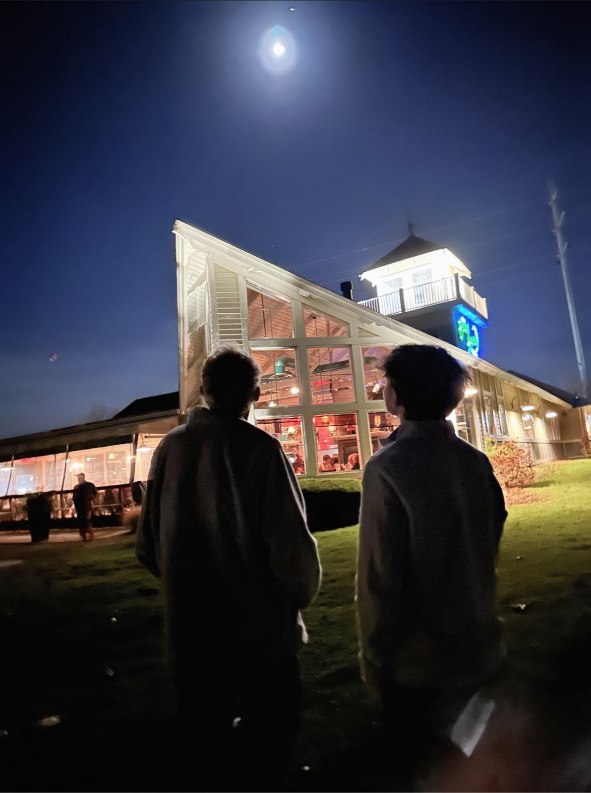 Two people standing on a lawn facing a brightly lit, steep-roofed building beneath the moon