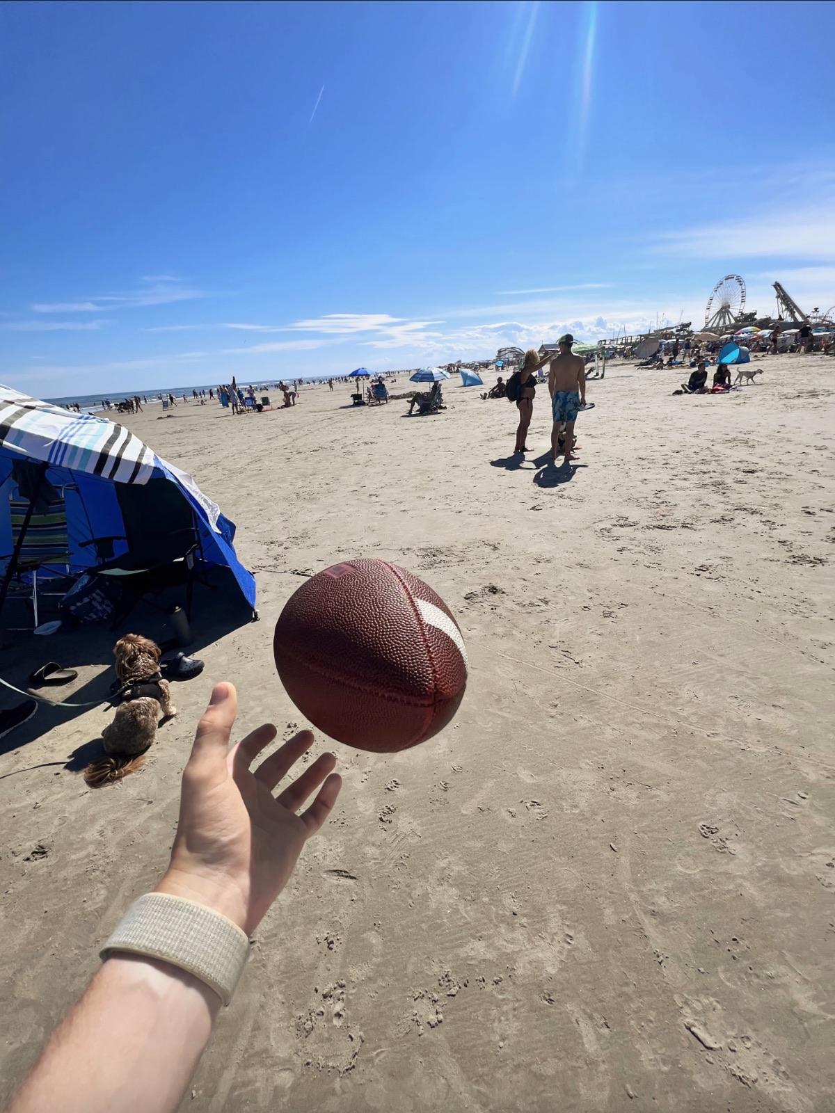 Hand tossing a brown football above a sandy beach with beachgoers, umbrellas, a dog, and distant amusement rides