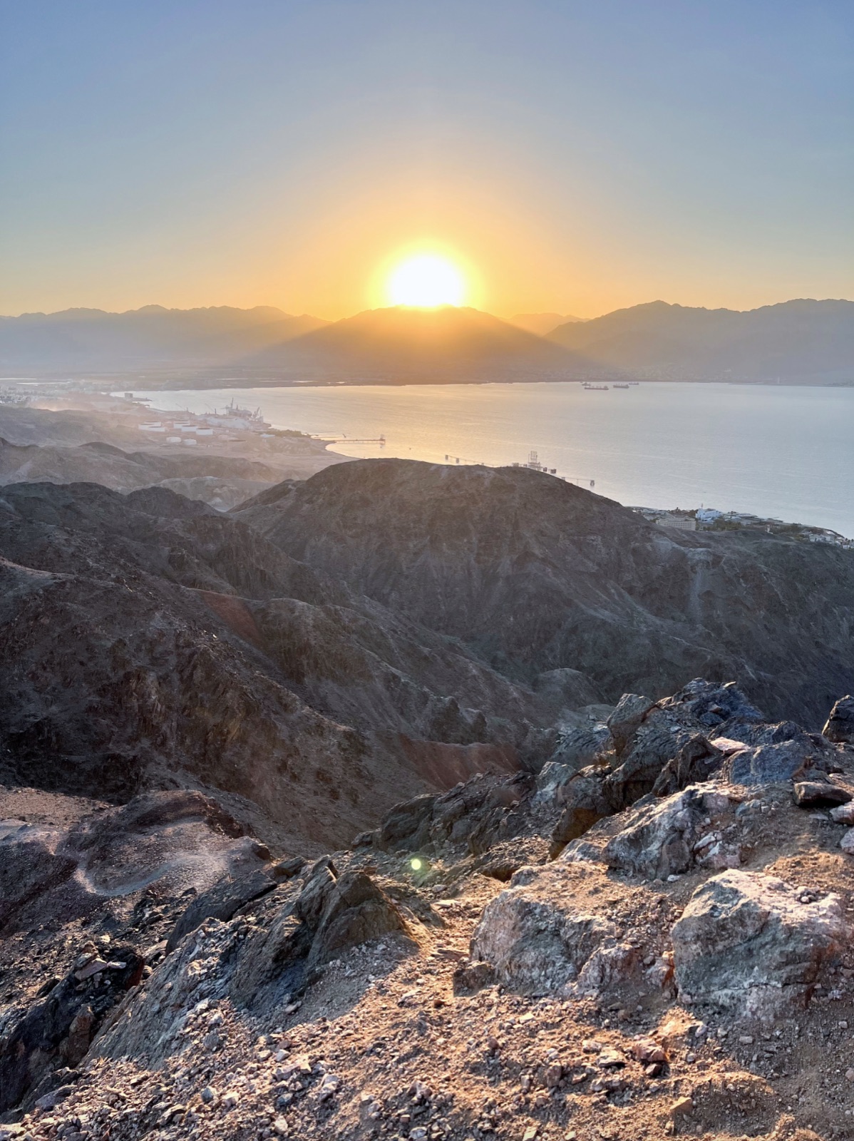 Low sun glowing above mountain ridges across a bay, with rocky hills and harbor facilities below
