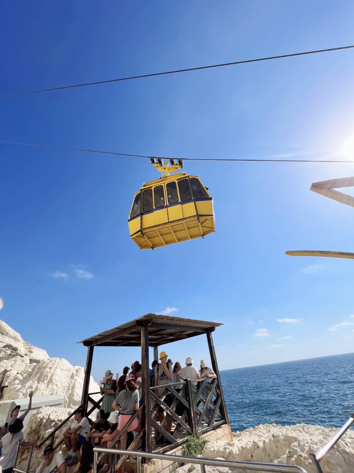 Yellow cable car suspended above people gathered at a rocky seaside platform