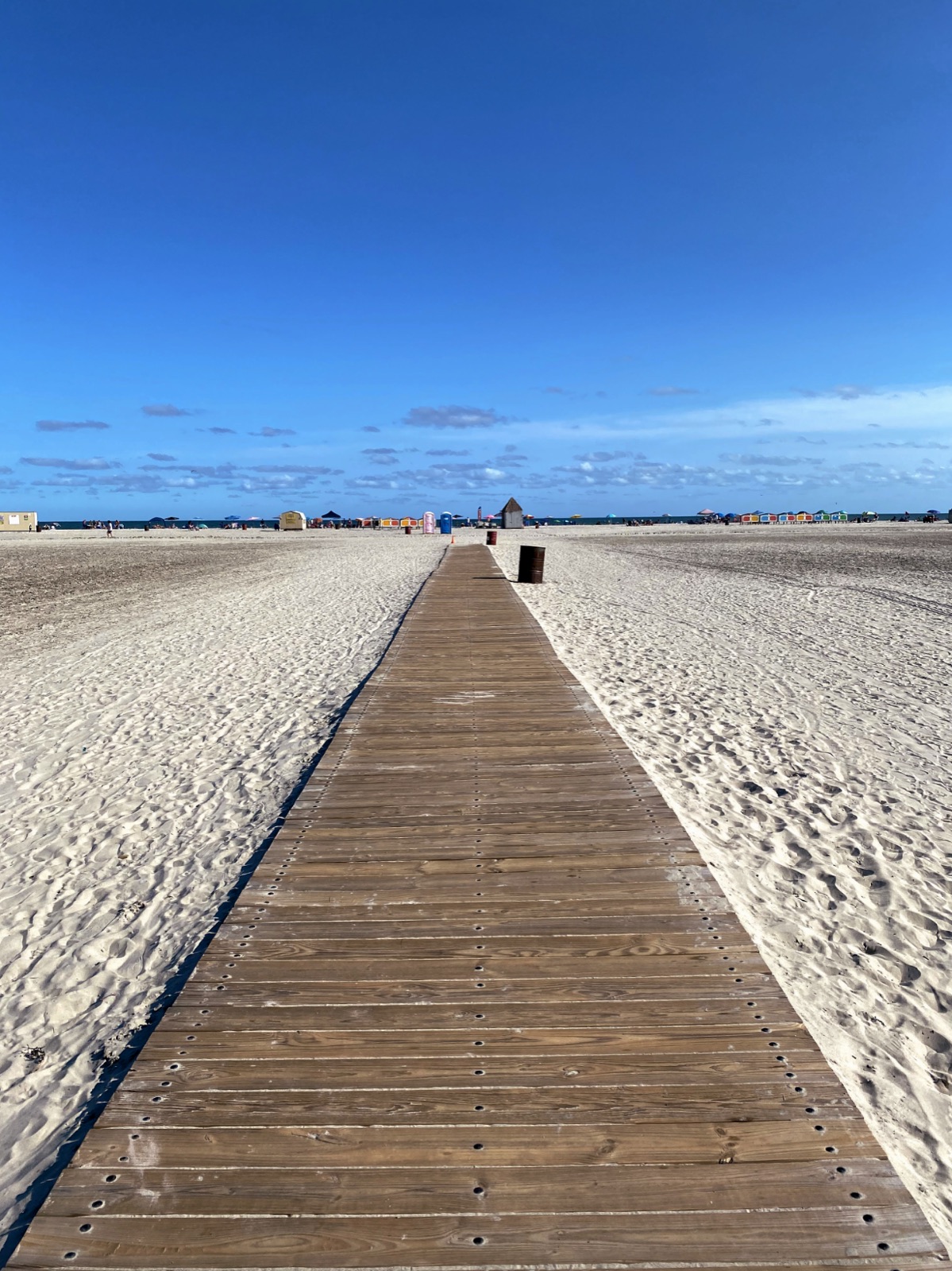 Long wooden walkway crossing pale sand toward colorful beach huts and the ocean
