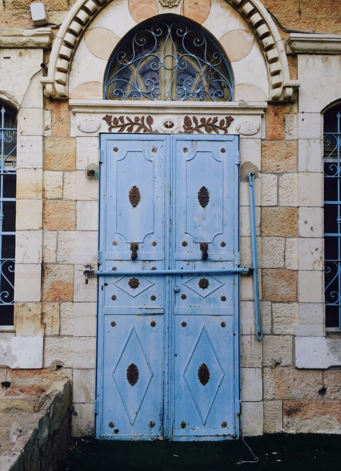 Weathered light-blue double doors with metal ornaments and an ornate arched transom in a stone facade