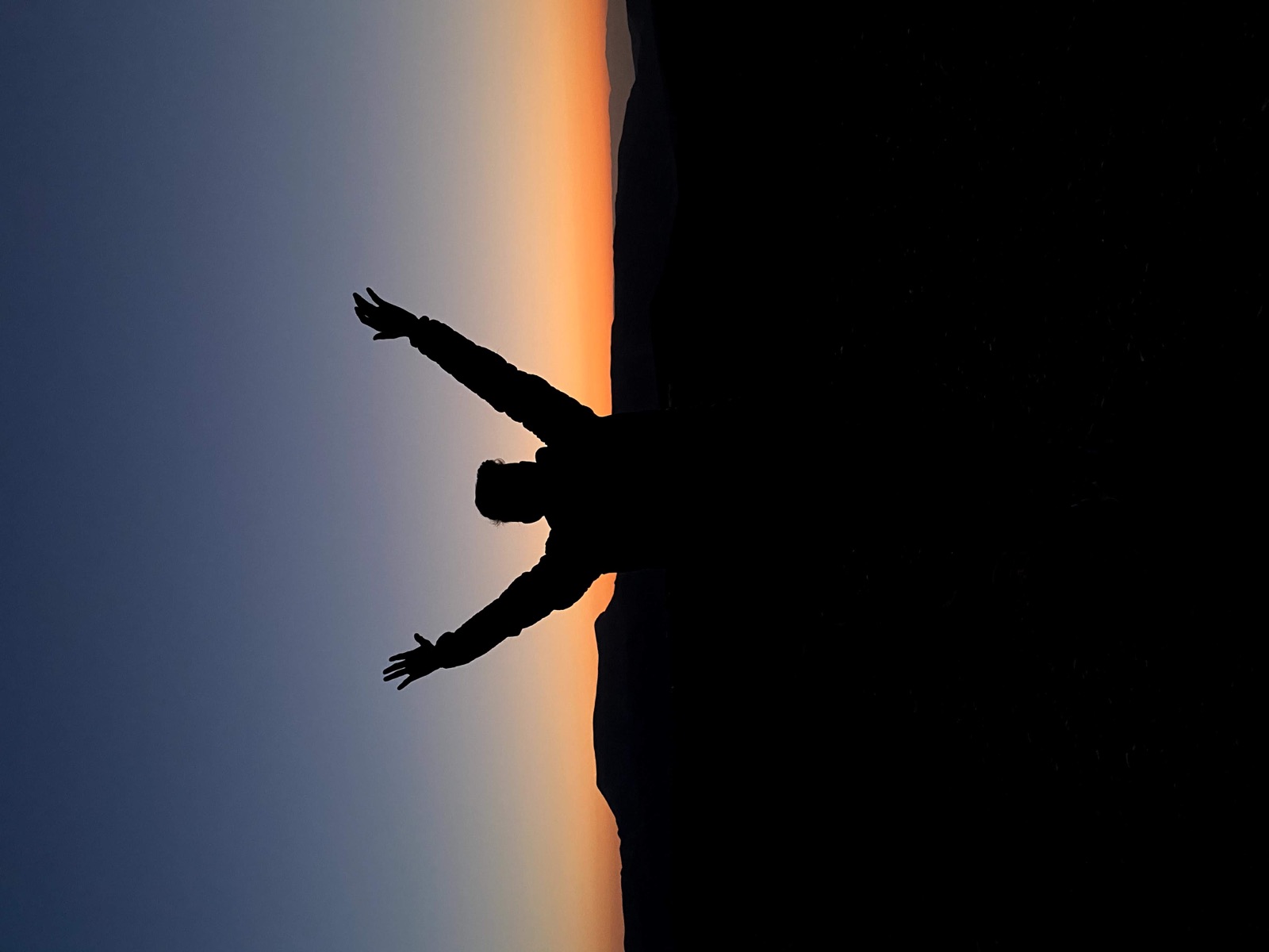 Silhouette of a person raising both arms against a sunset above distant hills